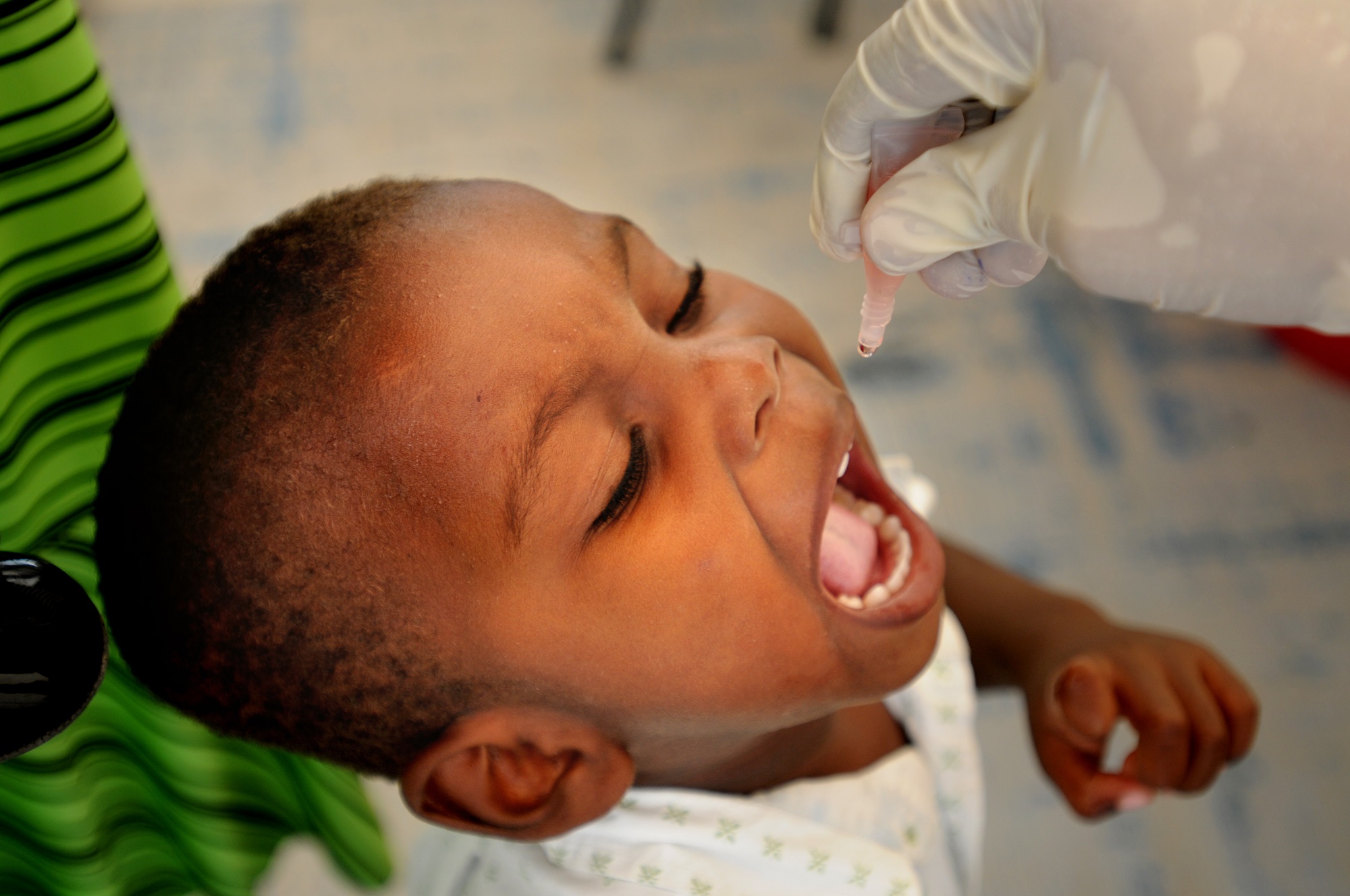 Menino de pele negra está com a boca aberta, recebendo uma dose de vacina em formato de gota. Uma mão com luva segura uma ampola sobre sua boca. Crédito: Kendra Helmer/USAID/Flickr. 