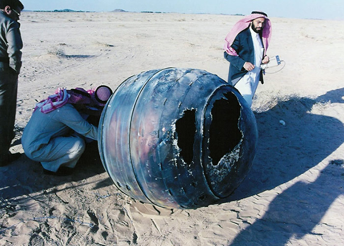 Homem com roupas típicas da Arábia Saudita observa, em meio ao deserto, grande tambor de metal com marcas de combustão. Outro homem está abaixado, na altura do chão, e manipula este tambor. (Crédito: Space Research Institute, King Abdulaziz City for Science and Technology, Riad, Arábia Saudita /Nasa)